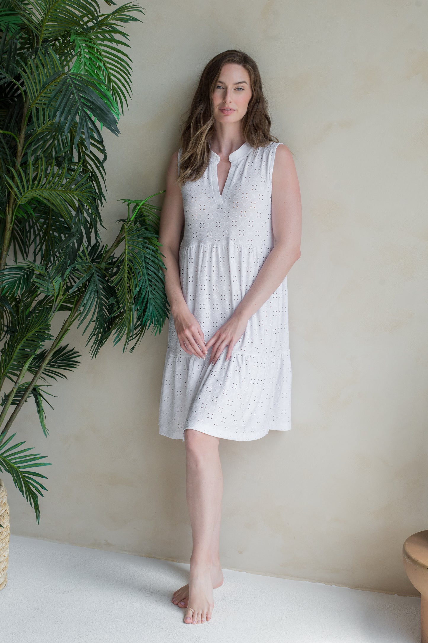 Woman in a white sleeveless dress standing next to a potted plant against a beige wall.