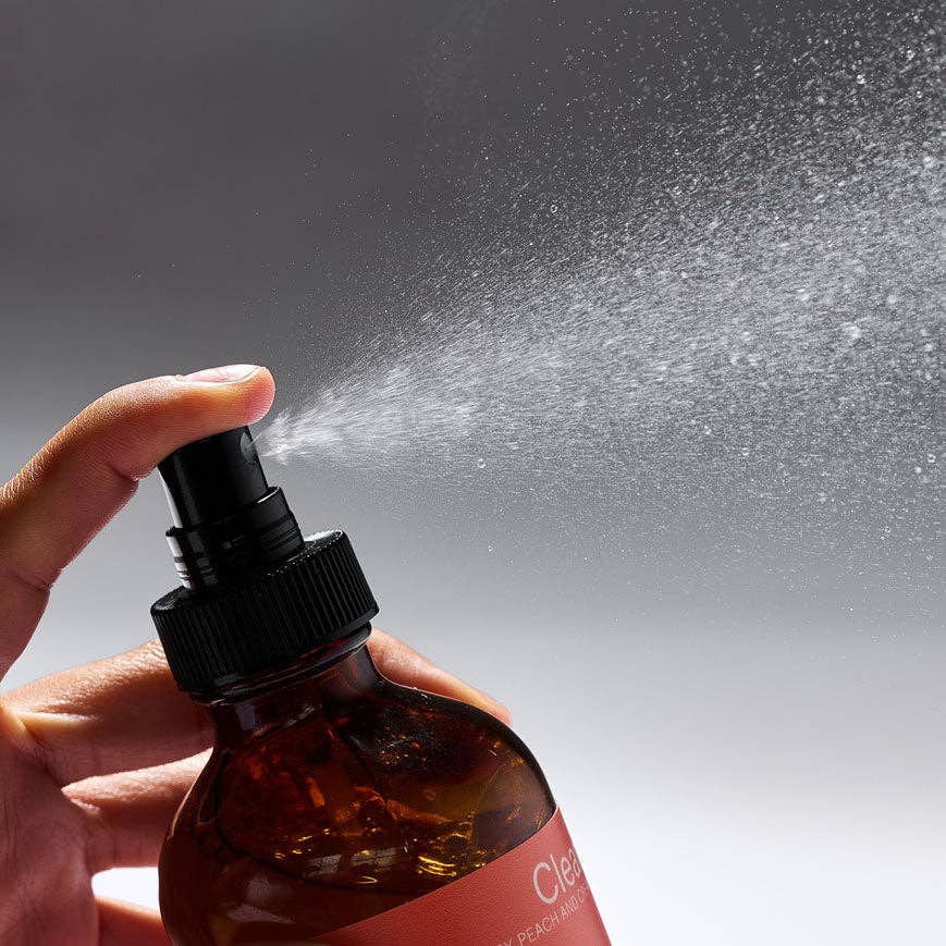 Hand holding a brown spray bottle with a orange label, spraying a mist against a gray background.