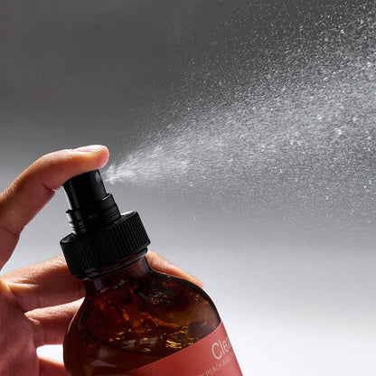 Hand holding a brown spray bottle with a orange label, spraying a mist against a gray background.