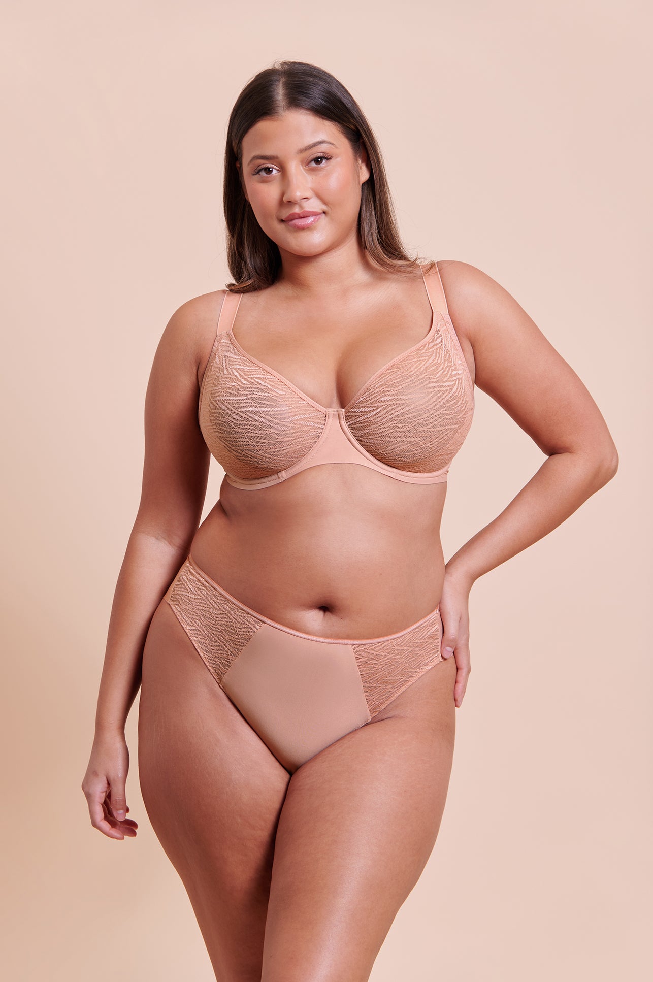 A model wearing a two-piece lingerie set in a light peach-beige color, featuring a lace underwire bra and matching lace high-waisted briefs, set against a solid beige studio background.